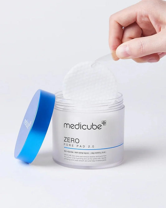 A hand lifting a white Medicube Zero Pore Pad 2.0 from a clear jar with a blue lid.